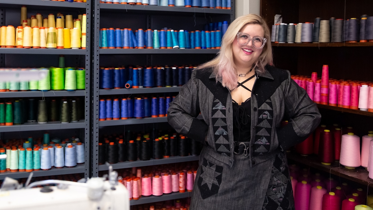 Mary Nease stands in front of a row of spools of thread, which are organized by color.