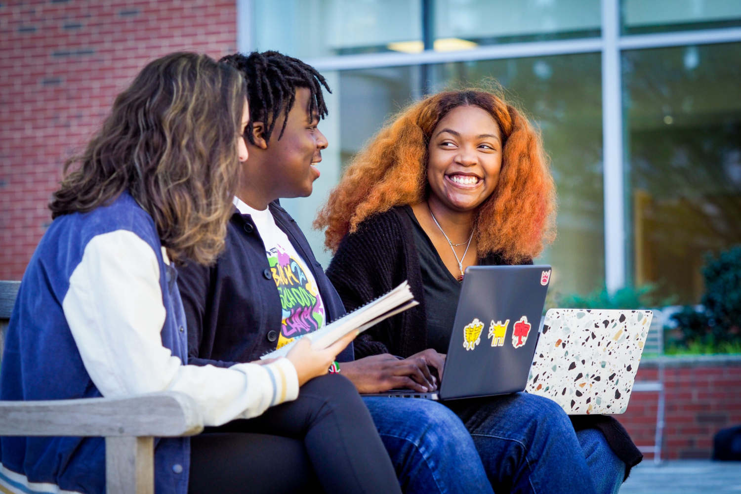 Three students sit outside on NC State's campus smiling with laptops in their laps.