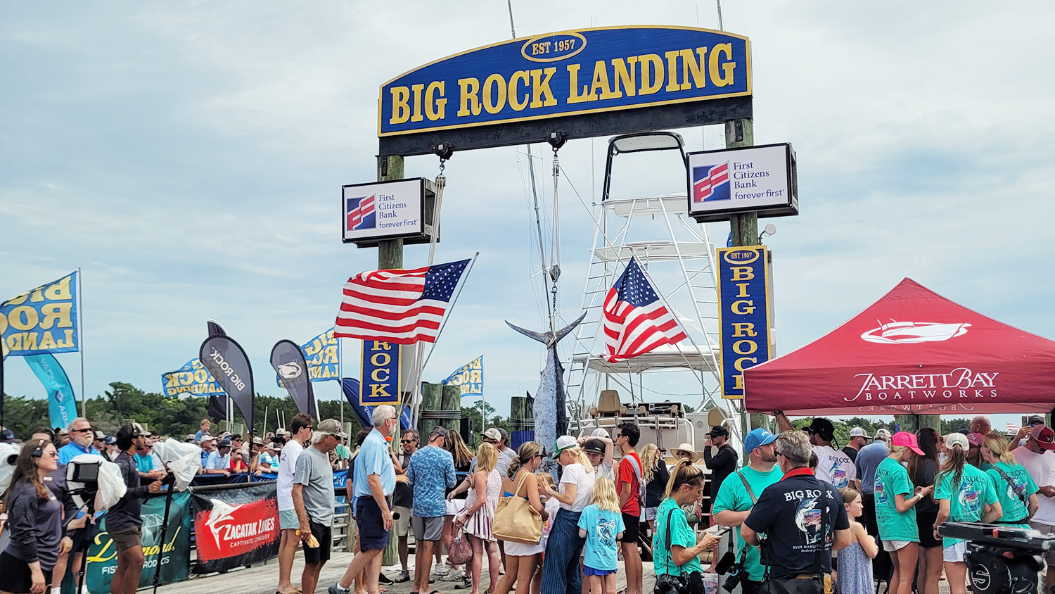 a crowd gathered near a dock and boats looking at a recently caught blue marlin