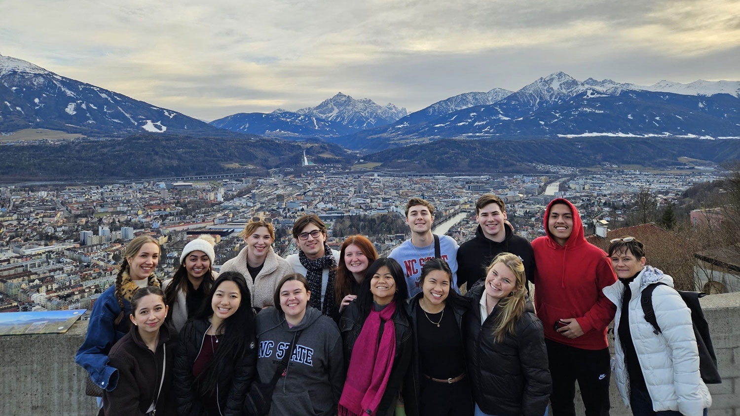 A group of students pose in front of a European cityscape with snowy mountains showing the background.
