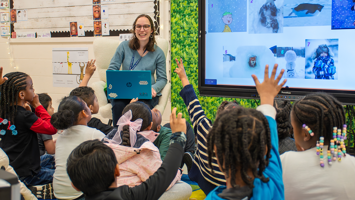 A school counselor sits at the front of a classroom in front of students, one of whom is raising their hand.
