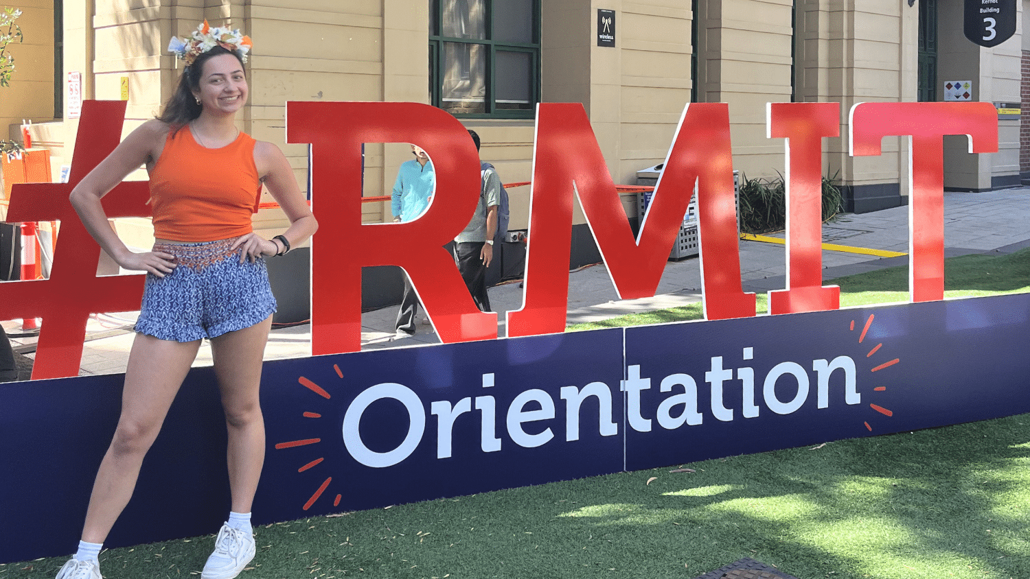 Annabelle Sharp poses with her hands on her hips and a smile in front of a sign reading "#RMIT Orientation" in large red letter. The signs spans the entire image and is on a lawn in front of tan buildings and a walkway.
