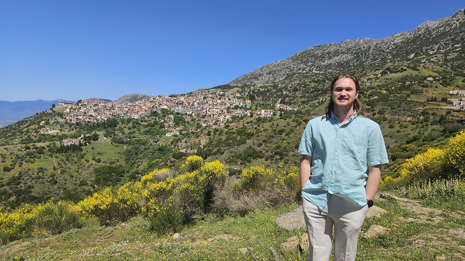 a man wearing a light teal shirt and khaki pants stands in front of a greek moutainside village