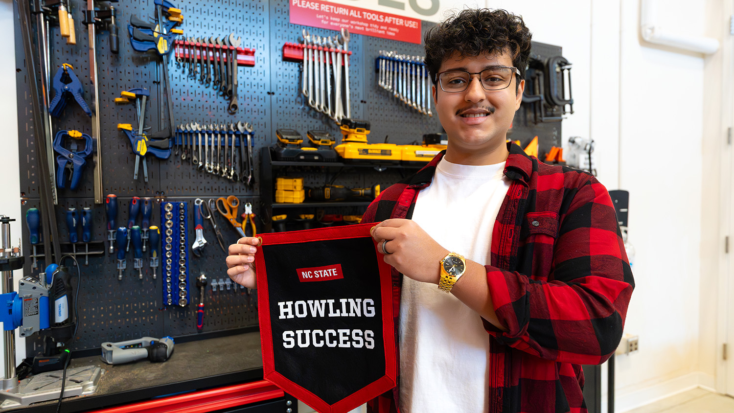 Daniel Briceno, the February Division of Academic and Student Affairs Howling Success, holds up his black and red Howling Success pennant in front of a work table and wall bearing several tools.