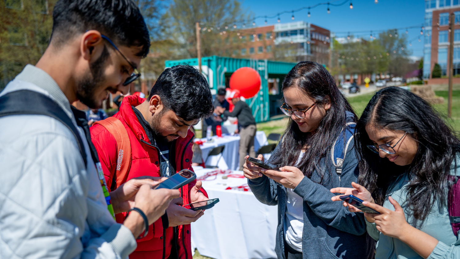 Four people stand together outside on Centennial Campus and look at their phones.