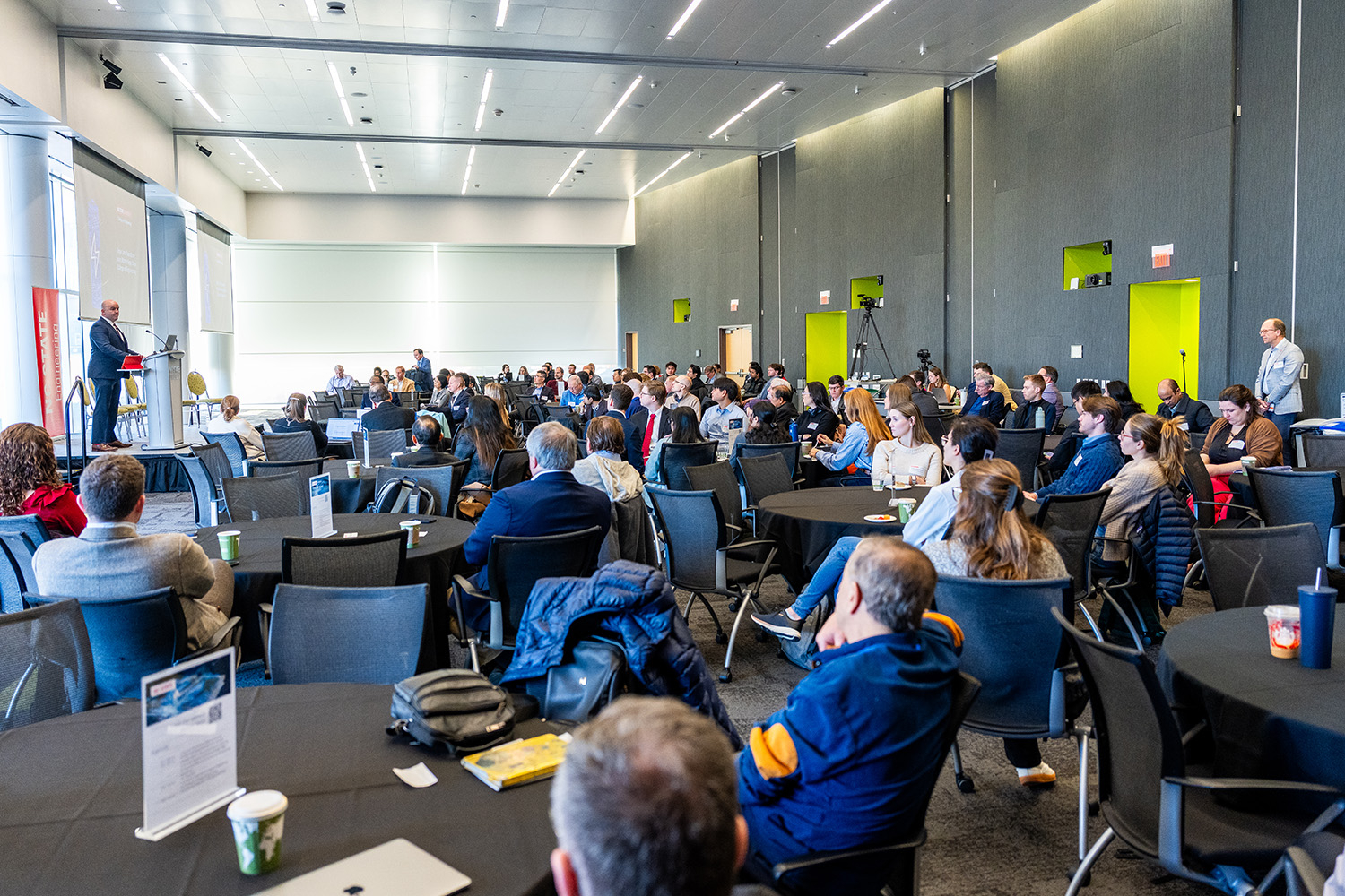 man standing behind a podium on an elevated platform, left, speaking to a large group of seated people in a long and open meeting space