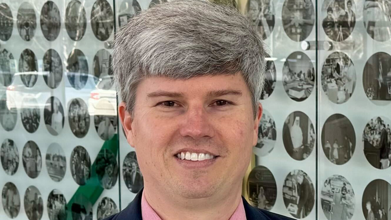 Gardinal Gibbons High School teacher and College of Education alum Taylor Blanton stands in front of a glass case with a series of photos behind him.
