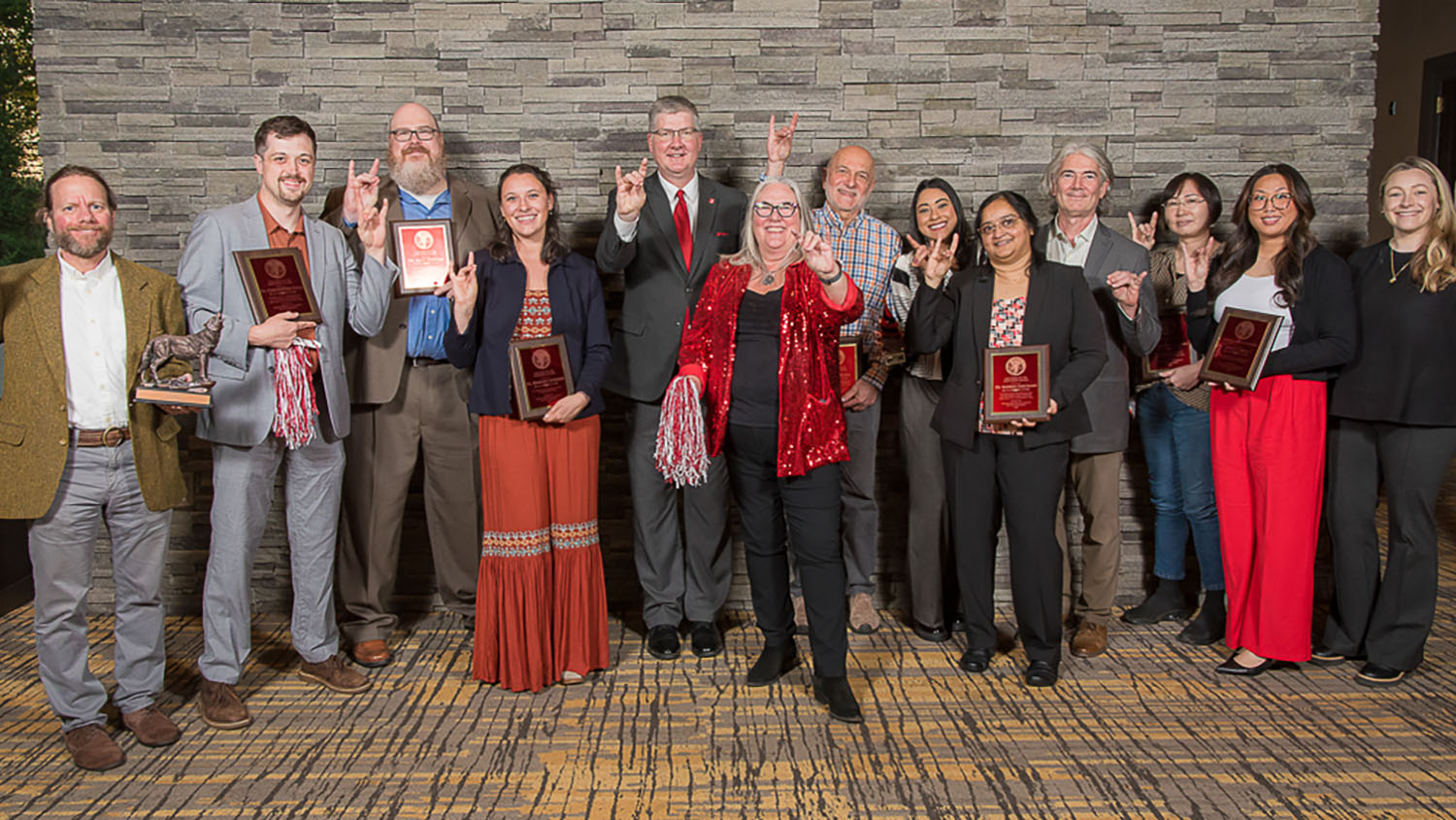 a group of people standing in front of a gray brick wall holding awards and making wolf ears signs with their hands