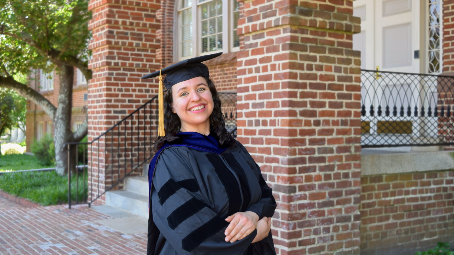 Ana Cubas Baez stands smiling in a graduation cap and gown in front of a brick building on NC State University's campus.