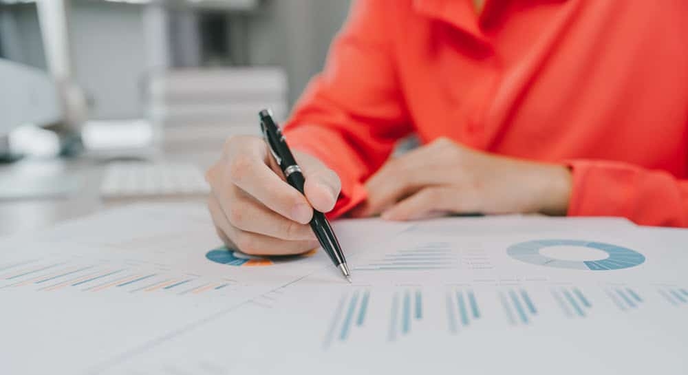 Person holding pen analyzing business report with charts and graphs on desk, wearing red shirt, focused on data review