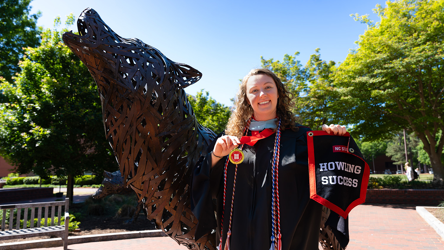 Marisa Rotolo, wearing her graduation gown, holds up her Howling Success banner in front of the Wolf Plaza statues.
