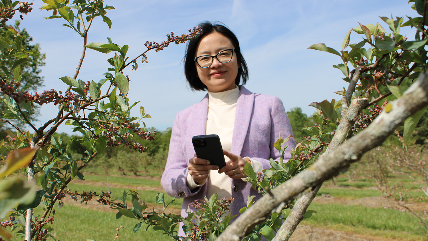 A woman wearing glasses and a lavender jacket holding a phone smiles at the camera through the branches of a bush.