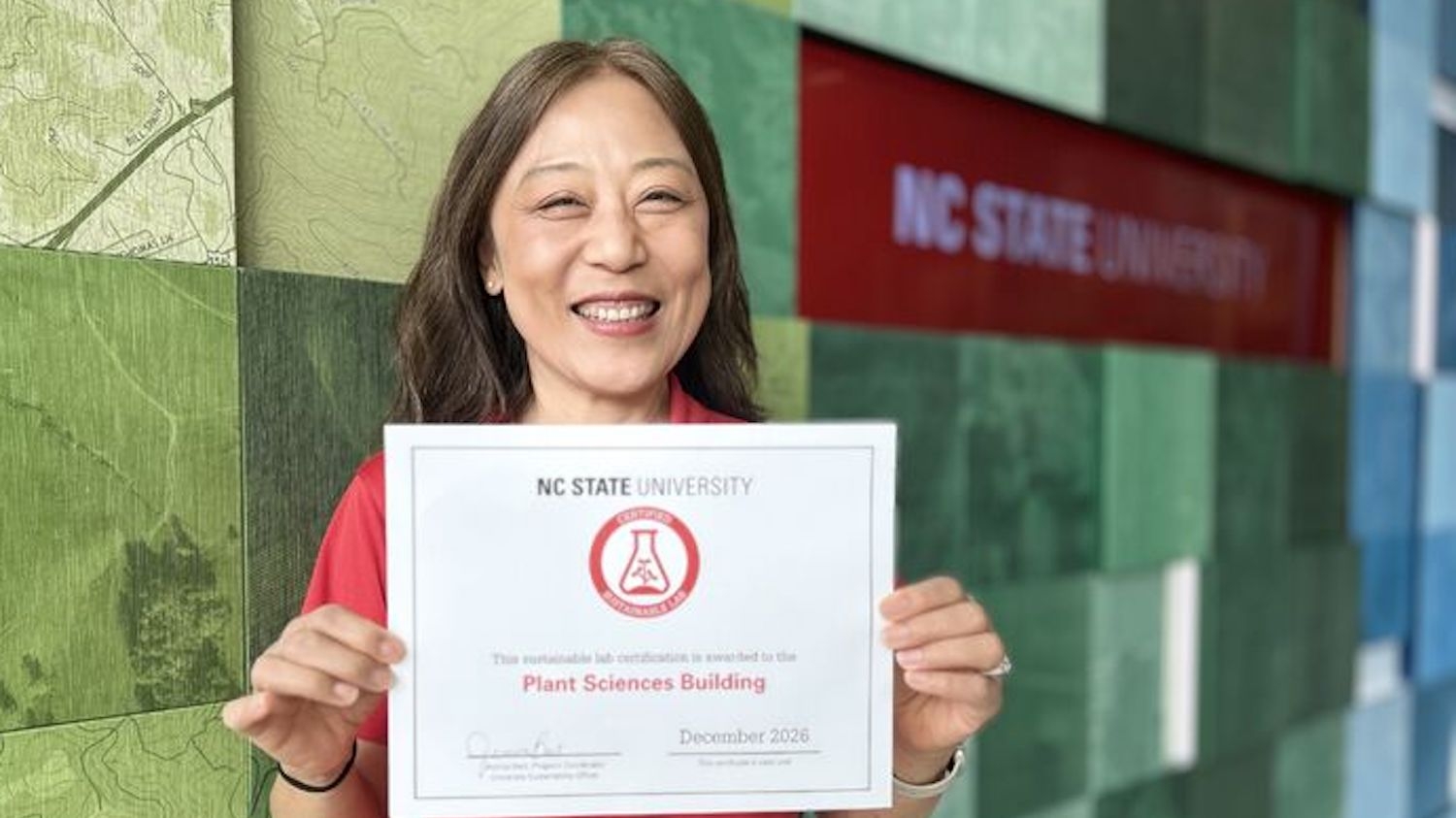 woman holds a certificate in front of a wall with the NC State University logo