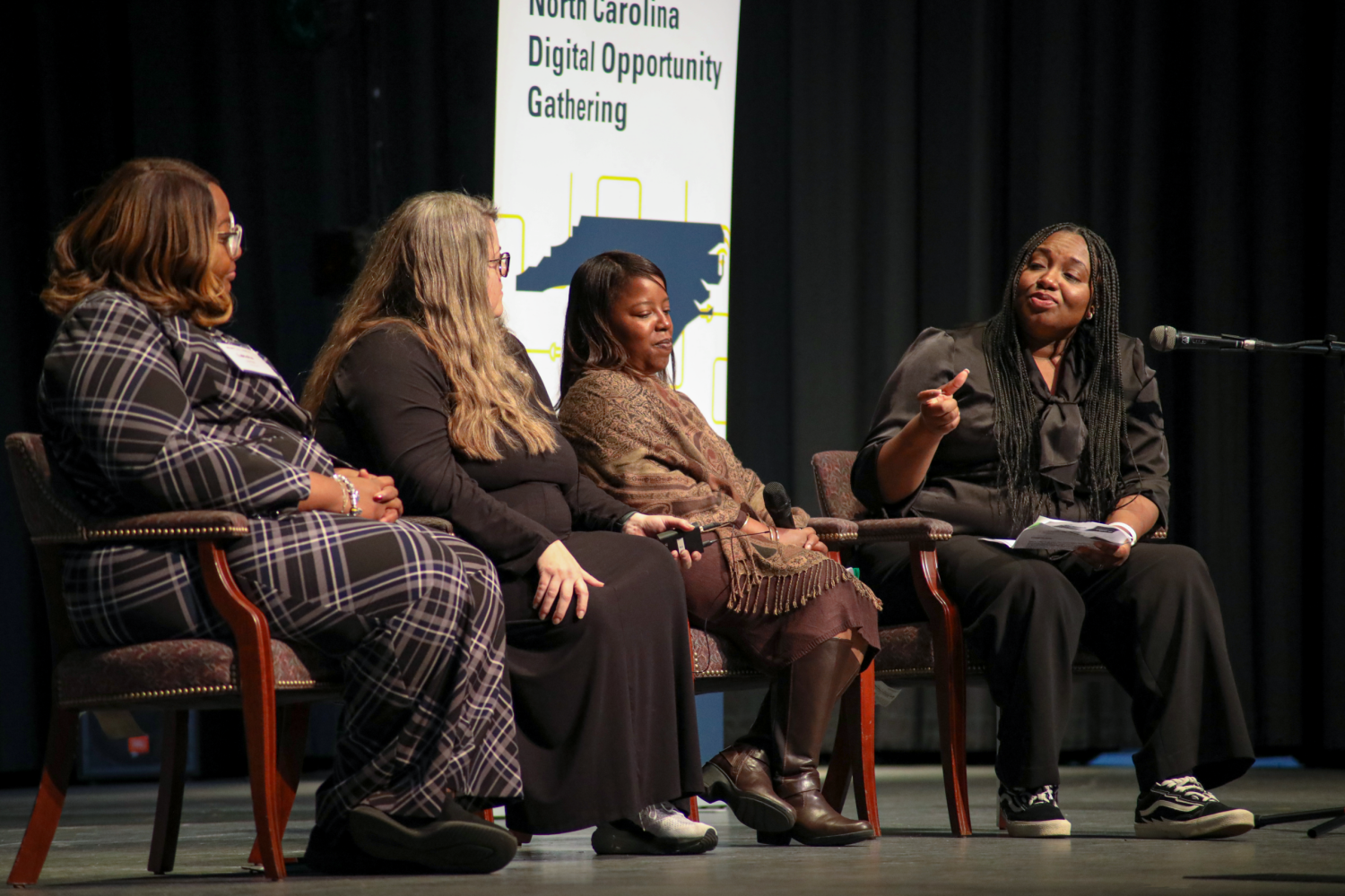 A line of four professionals sit on a stage in chairs in front of the NC Digital Opportunity Gathering standing banner