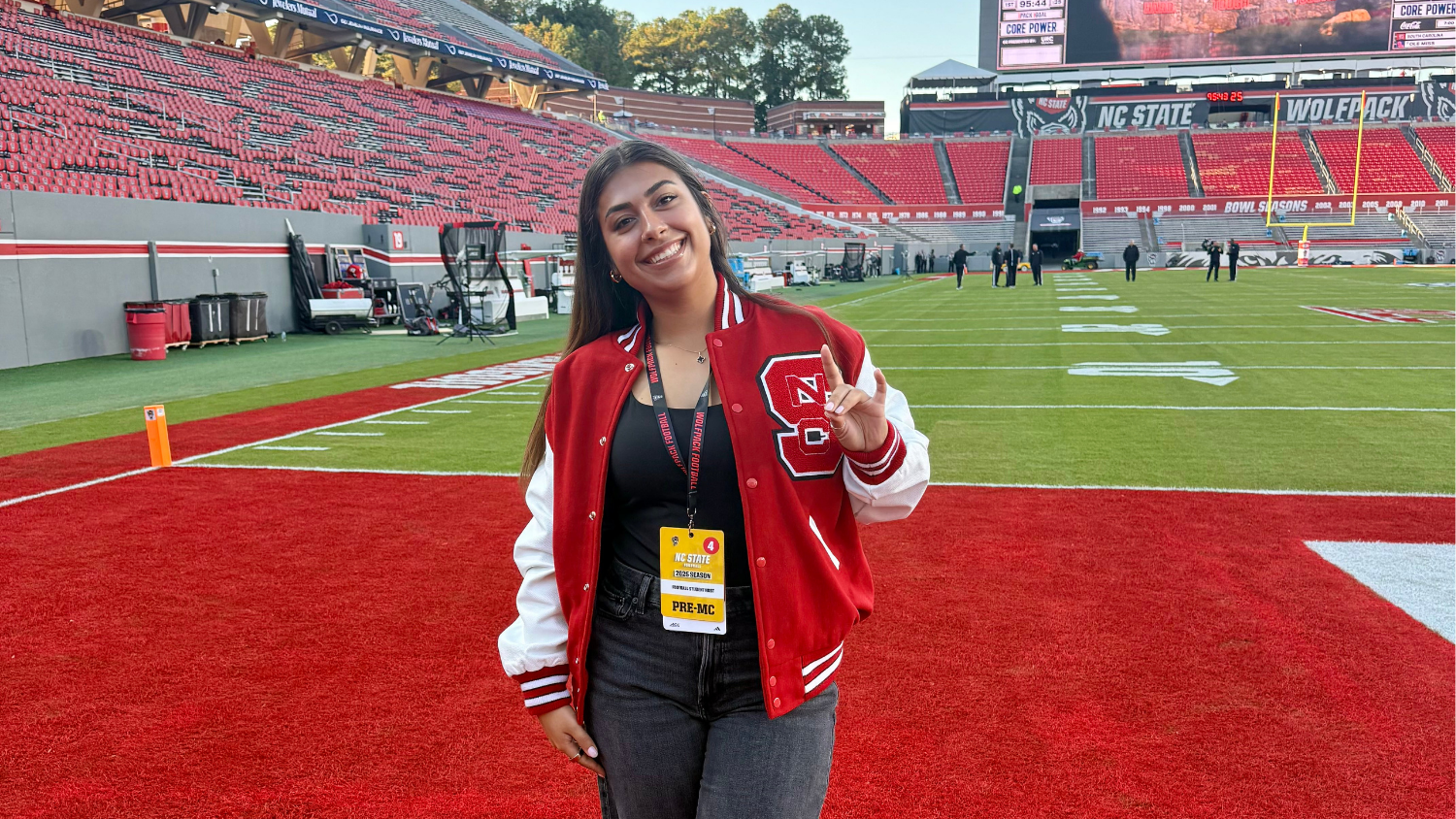 A young woman in a red varsity jacket smiles and gestures on a football field in front of empty red stadium seats.
