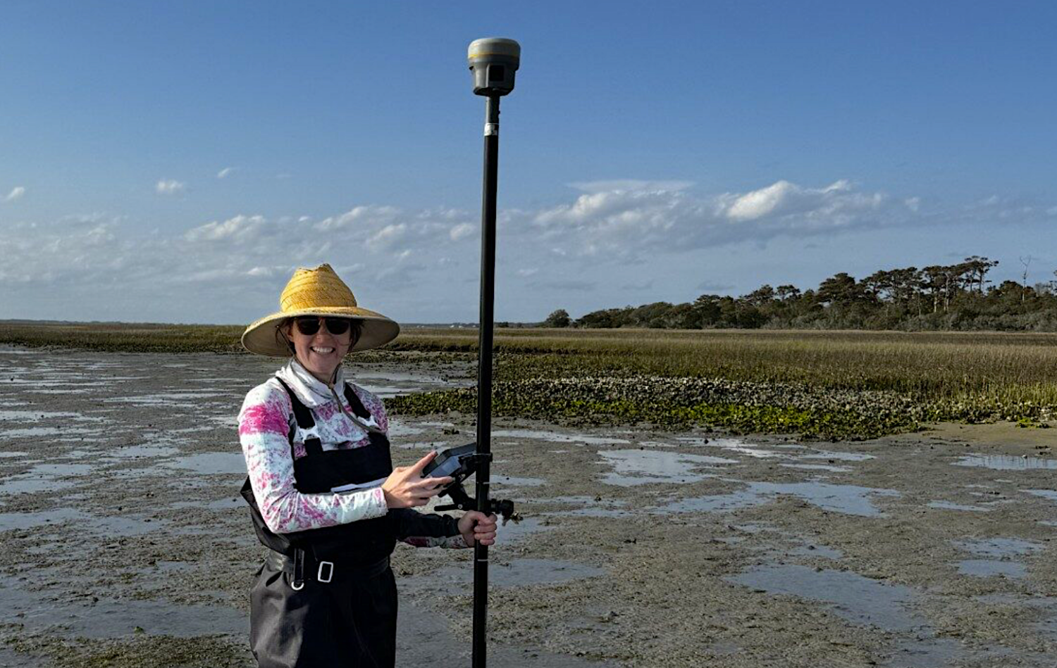 Woman wearing a hat and coveralls stands in a sea grass meadow with a tall scientific instrument.
