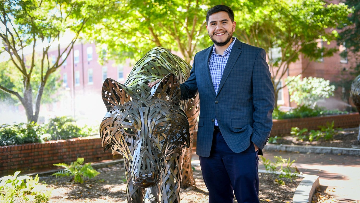 man with short dark hair and beard, wearing a dark blue suit and light blue shirt, stands next to a bronze statue of a wolf under shade trees