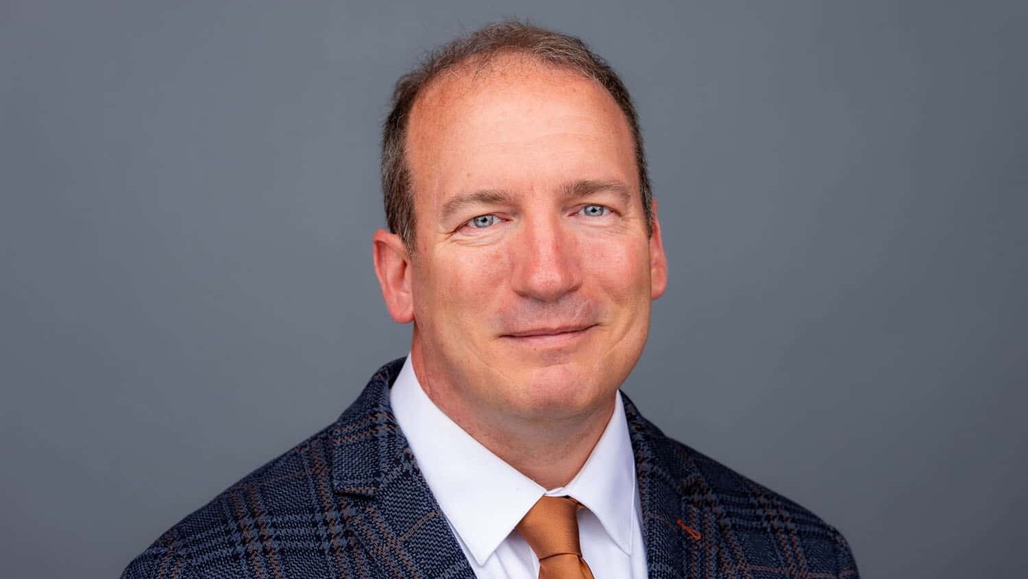 older man wearing a dark patterned sport jacket, white shirt and orange necktie