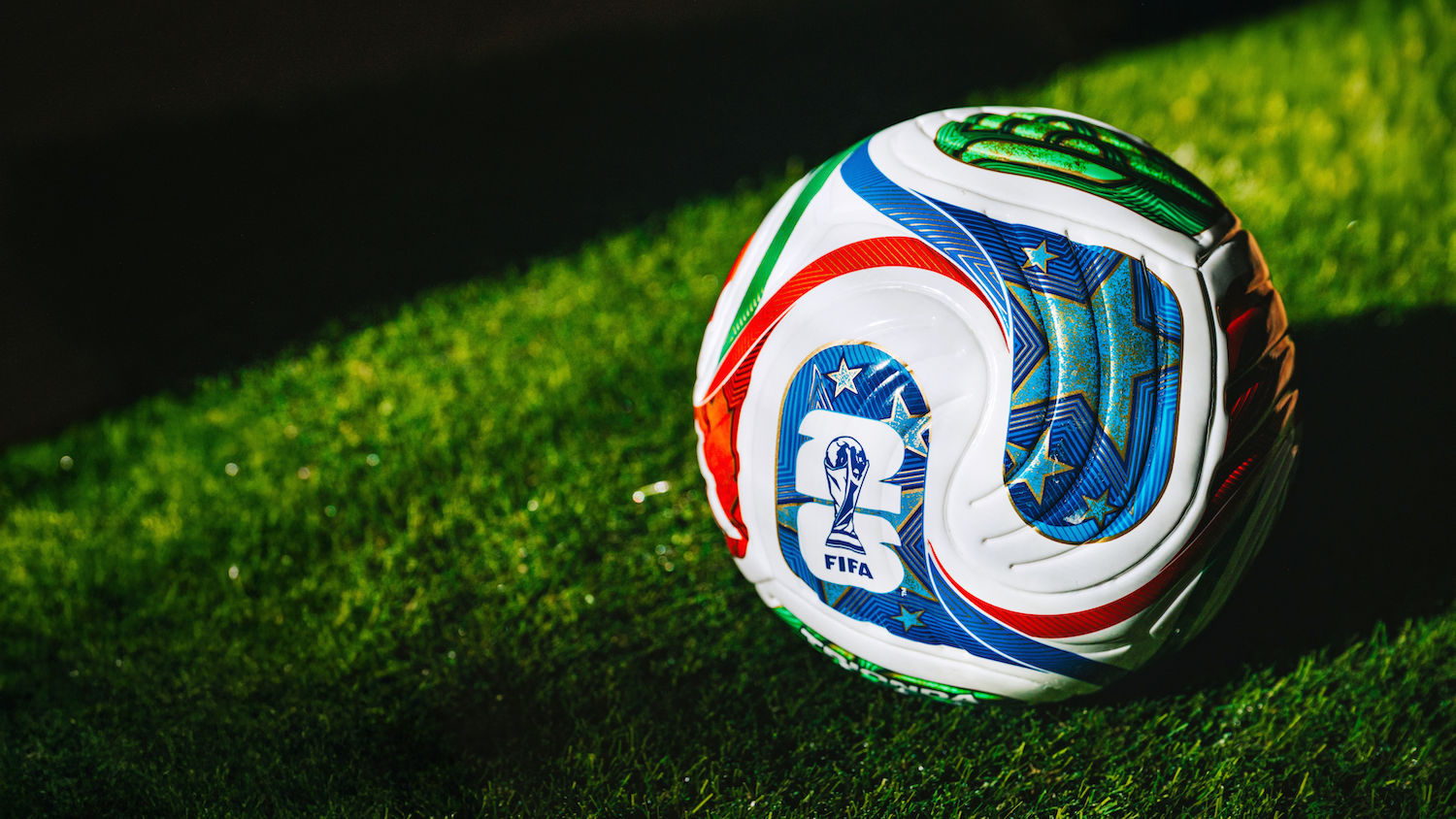 A colorful soccer ball with a FIFA emblem sits on green grass, partially lit against a dark background.