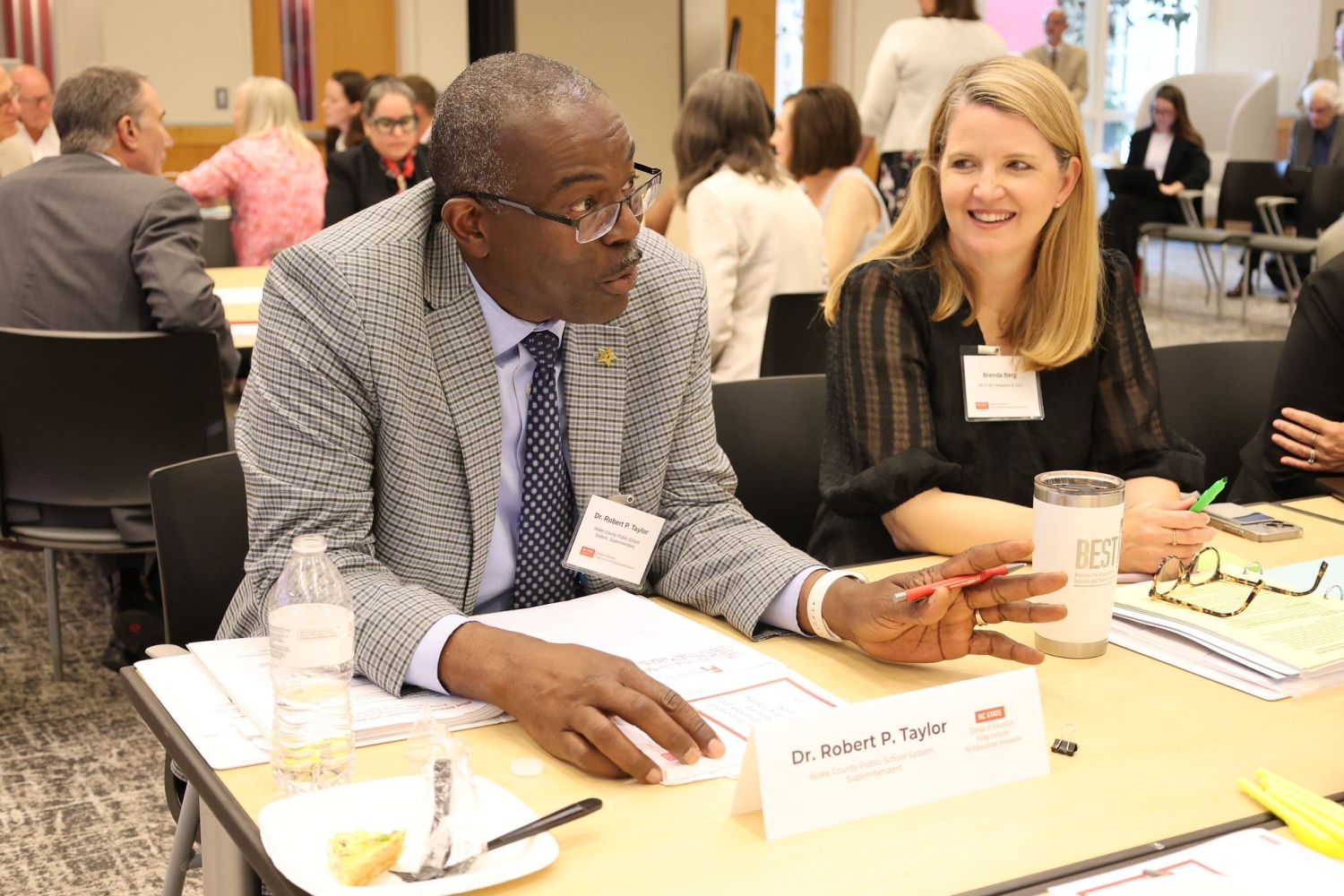 A man and woman have a discussion at a table of people. The man speaks with his hands while the woman smiles at him.