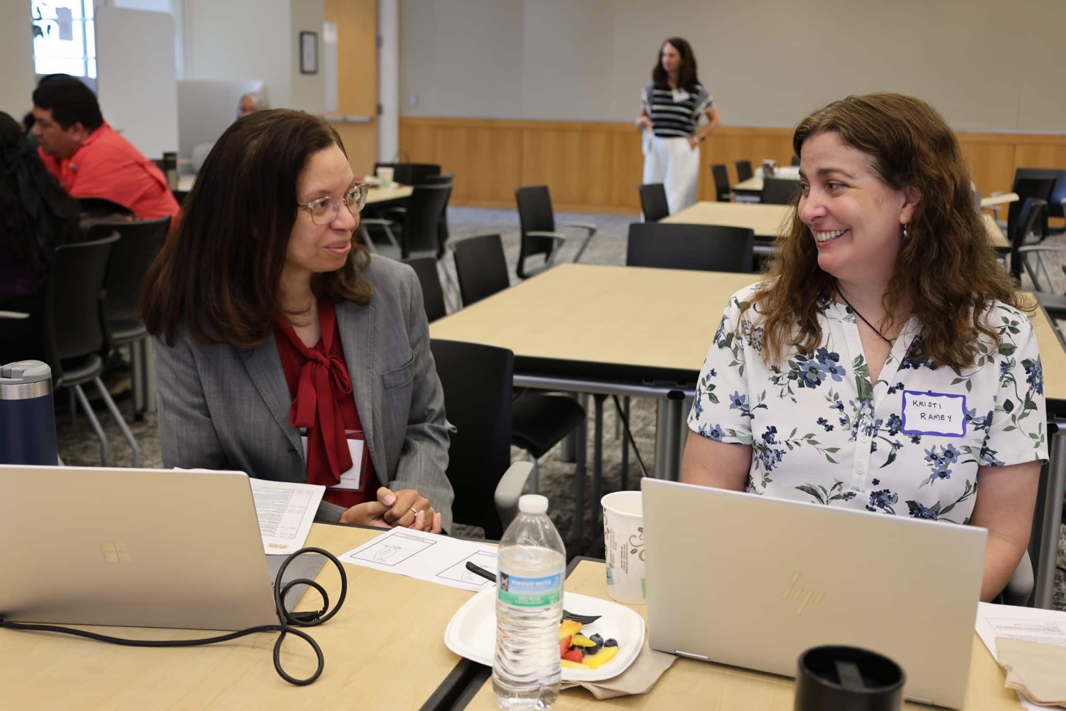 Two women sit next to each other at a table while talking and smiling. Laptops are in front of each of them.