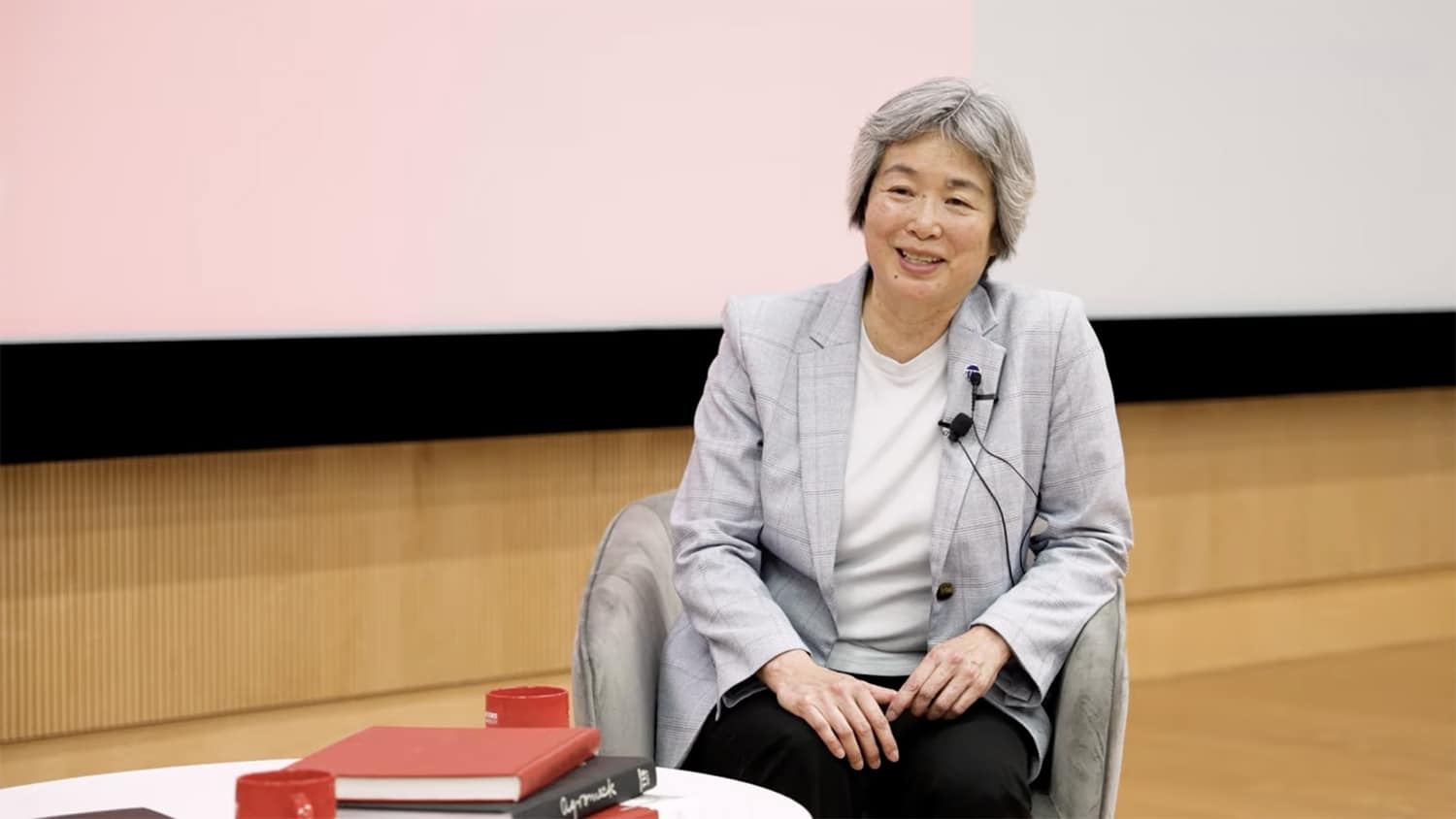 older woman with short gray hair, wearing a light grey jacket, white top and black pants; sitting in a chair with a small table in front of her