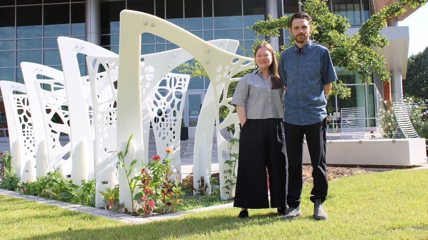 Two people standing side by side next to a multipanel outdoor trellis made of bright white plastic.
