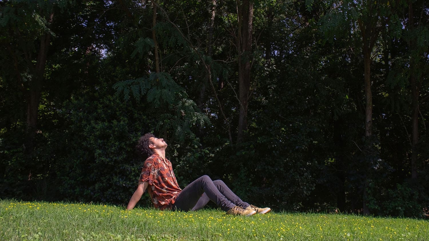 a young man lies back on the grass in a wooded area with his eyes closed, apparently listening to his surroundings