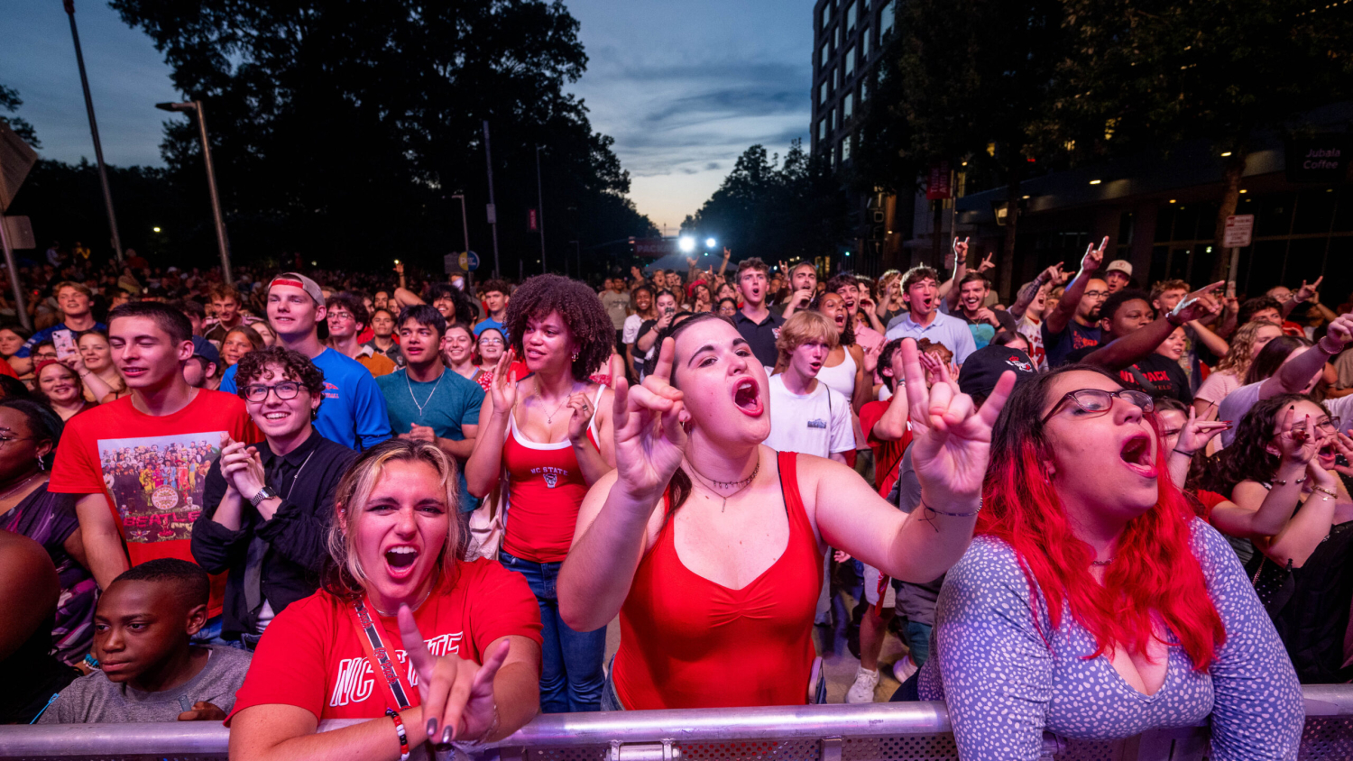 A lively crowd at an outdoor concert, cheering and raising their hands as the sun sets behind them.