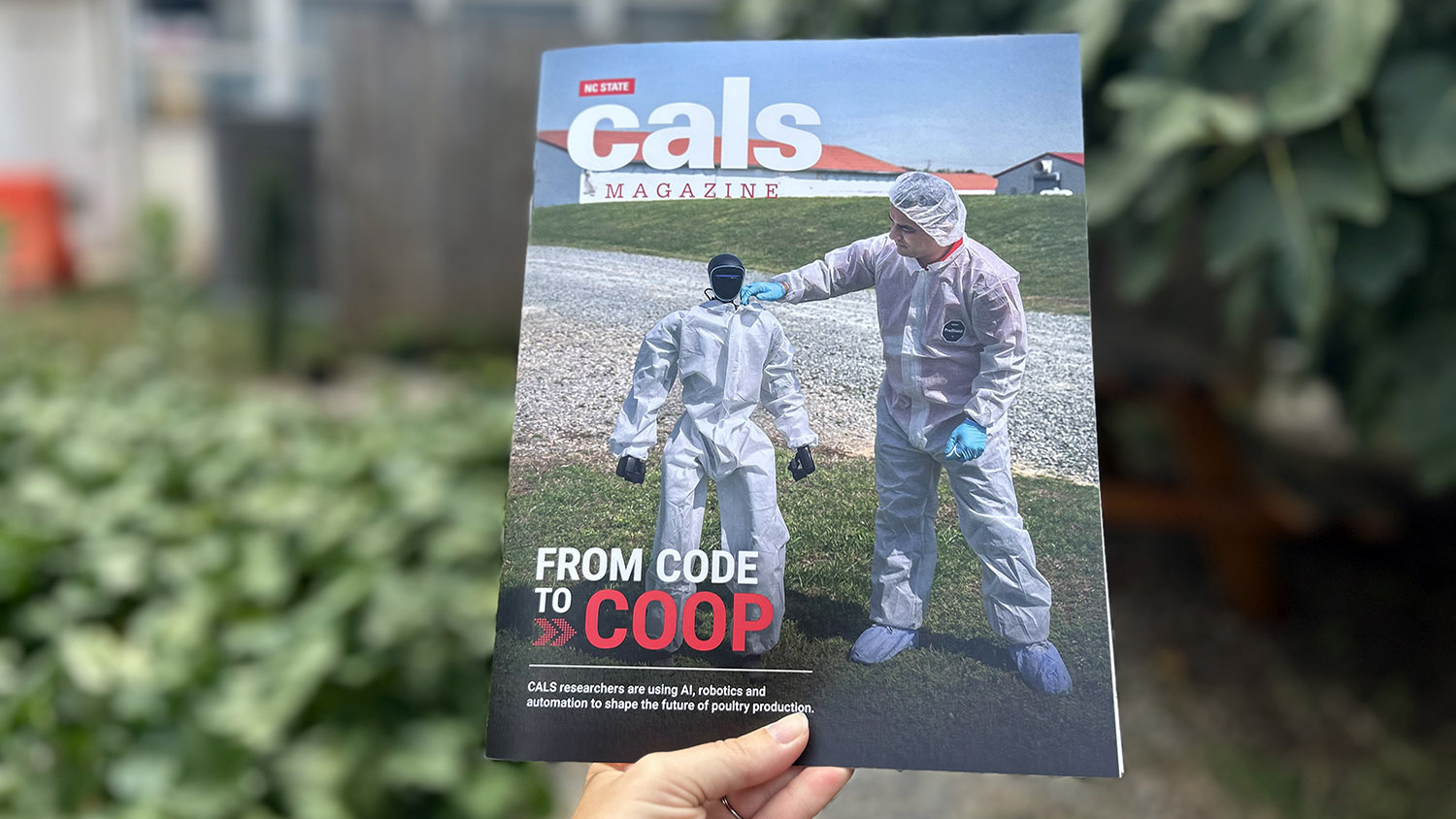 a person holds a magazine with an image of a humanoid robot and a man both in white coveralls in front of a building with a red roof and white walls