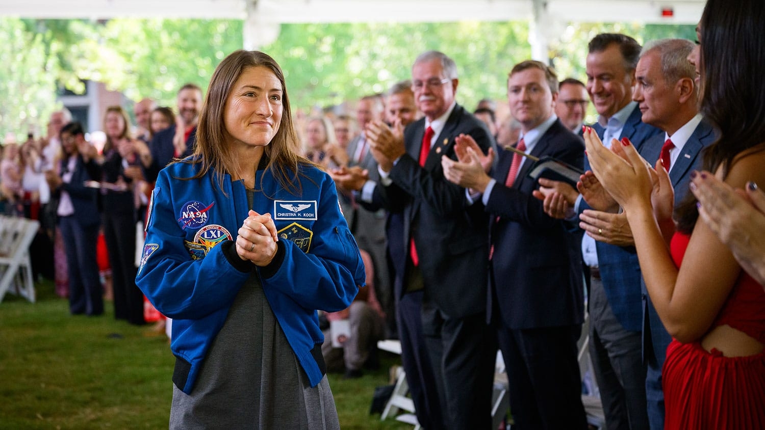 A person in a NASA flight jacket stands at an outdoor event, clapping as others around them applaud.