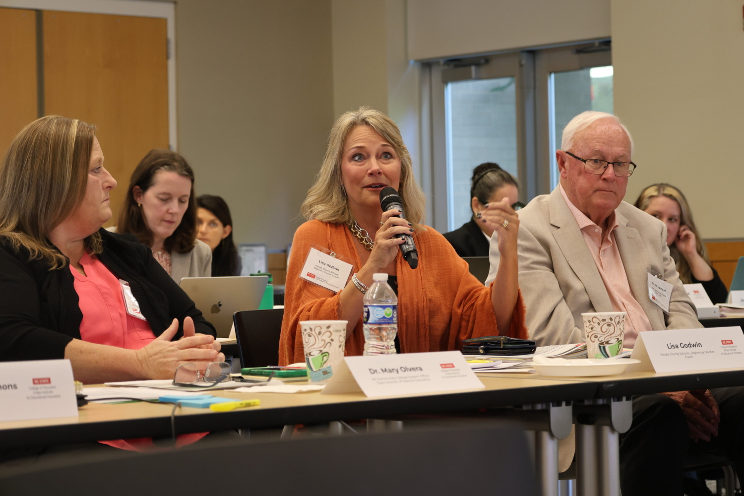 Commission member Lisa Godwin, a woman in an orange shirt, speaks into a microphone while sitting at a table.